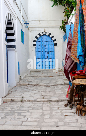 Sidi Bou Said. La Tunisia. Vista dei tappeti di souvenir in vendita lungo stretti vicoli in ciottoli di bianco e nero con blu arcuata in corrispondenza della porta Foto Stock