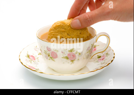 Mano caucasica femmina che cava un biscotto in una tazza di tè in una tazza di porcellana con piattino. Foto Stock