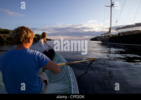 Un ragazzo e una ragazza riga in una Peapod dinghy al tramonto verso un classico yacht ormeggiati lungo la costa del Maine. Foto Stock