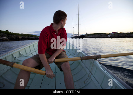 Un giovane uomo di righe in una Peapod dinghy al tramonto verso un classico yacht ormeggiati lungo la costa del Maine nei pressi di Boothbay Harbor. Foto Stock