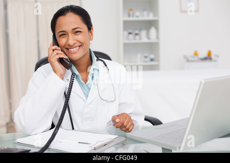 Medico sorridente a parlare al telefono mentre è seduto nel suo ufficio Foto Stock