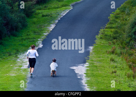 Una madre corre a fianco di suo figlio a cavallo di un equilibrio bici giù per una ripida collina. Foto Stock