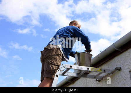 Uomo su una scala la pulizia delle grondaie su un cottage Foto Stock