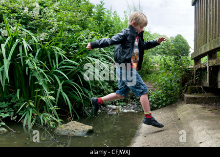 8 anno vecchio ragazzo caucasico saltando su rocce nel flusso a barre corte Moat stagno, vicino Longwell verde in Bristol, Regno Unito Foto Stock