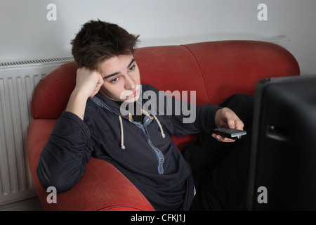 Giovane uomo guardando la TV, tenendo lo stesso telecomando, guardando annoiato. Foto Stock