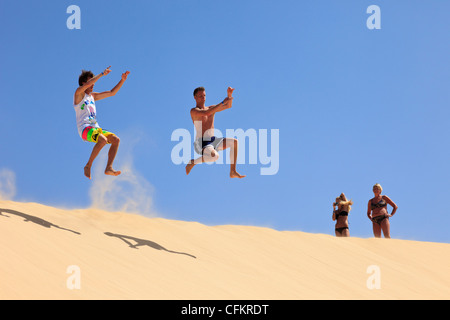 Due uomini che saltano in aria dalla cima di una grande duna di sabbia con due donne che guardano e scattano foto in una giornata di sole. Boa Vista, Isole di Capo Verde Foto Stock