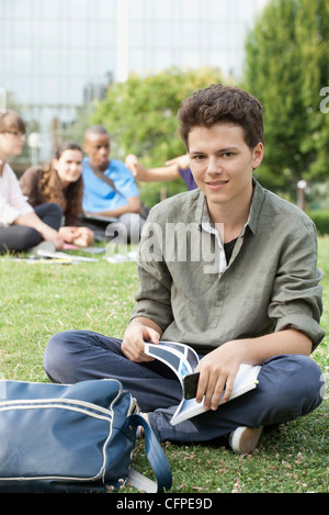 Giovane uomo seduto su erba, persone in background, ritratto Foto Stock