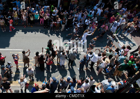 La Torcia olimpica arriva giù Coldharbor Lane in Brixton, Londra, Regno Unito. I torchbearer è protetto dalla polizia in grigio sportswear e il relè è preceduta da sponsor veicoli. Il 26 luglio 2012. Foto Stock