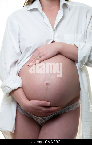 Una giovane donna incinta mentre tiene il suo pancino indossa pantaloni bianchi e un mans camicia bianca su sfondo bianco Foto Stock