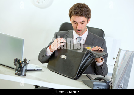 Scioccato imprenditore moderno seduto alla scrivania in ufficio e guardando in borsa Foto Stock