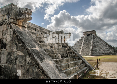Chichen Itza Tempio di Kukulkan Venere piattaforma Giaguaro Capi Messico // CHICHEN ITZA, Messico - teste giaguaro scolpite della piattaforma Venere occupano il primo piano nella zona archeologica di Chichen Itza, con il Tempio di Kukulkan (noto anche come El Castillo) visibile sullo sfondo. La piattaforma Venere, una delle numerose strutture cerimoniali dell'antico sito Maya, presenta intricate incisioni in pietra che riflettono la sofisticata conoscenza astronomica e le pratiche religiose della civiltà. Il Tempio di Kukulkan, una piramide a gradini dedicata alla divinità serpente piumata, sorge di circa 30 mete Foto Stock