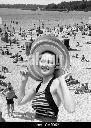 1930s VACANZA MONTAGE ritratto donna sorridente IN COSTUME DA BAGNO indossando grande cappello di paglia e la scena della spiaggia affollata Foto Stock