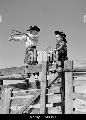 Anni Cinquanta due giovani ragazzi vestito da cowboy SLINGSHOTS RIPRESA SULLA PARTE SUPERIORE DELLA RECINZIONE DI LEGNO PER ESTERNO Foto Stock
