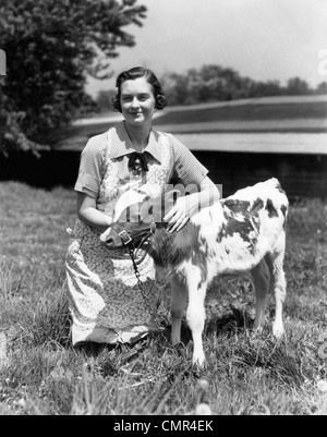 Negli anni quaranta anni cinquanta FARM DONNA IN grembiule inginocchiato in erba con il giovane vitello JERSEY Foto Stock