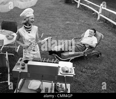 Anni Cinquanta anni sessanta la donna a chef del cappello e grembiule grigliare hot dogs sul grill UOMO IN CHAISE LONGUE a ridere di mangiare un FRANKFURTER AL DI FUORI Foto Stock