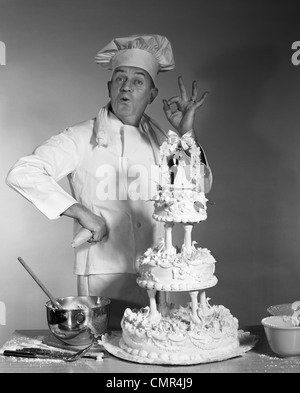 Anni Cinquanta anni sessanta anni settanta uomo ritratto BAKER guardando la fotocamera facendo segno OK con la mano accanto a tre livelli la torta di nozze la sposa e lo sposo sulla parte superiore Foto Stock