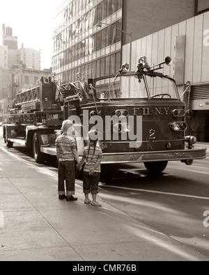 Anni Settanta 2 bambini ragazzo ragazza tenendo le mani guardando il camion dei pompieri parcheggiato sulla strada di NEW YORK CITY Foto Stock