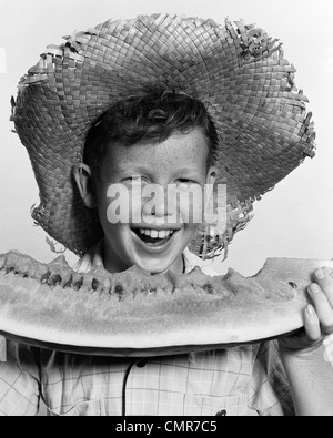 Negli anni quaranta anni cinquanta FRECKLE-di fronte ragazzo indossa cappello di paglia mangiando grandi zeppa di anguria guardando la fotocamera Foto Stock