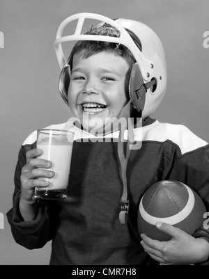 Anni sessanta sorridente giovane ragazzo con i baffi DI LATTE IN UNIFORME DI CALCIO & casco palla sotto braccio che tiene un bicchiere di latte guardando la fotocamera Foto Stock