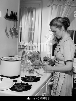 Anni Cinquanta il bambino nel seggiolone GUARDANDO LA MADRE IN CUCINA STUFA A PREPARARE IL LATTE PER IL BAMBINO IN BOTTIGLIA Foto Stock