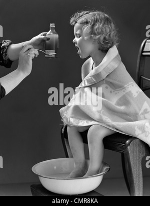 Negli anni quaranta malati bambina seduti in poltrona avvolto in una coperta piedi ammollo in acqua calda di prendere la medicina sul cucchiaio da madre Foto Stock