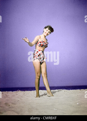 Negli anni quaranta anni cinquanta sorridente giovane donna indossa PLAID IN DUE PEZZI COSTUME DA BAGNO permanente sulla spiaggia di sabbia