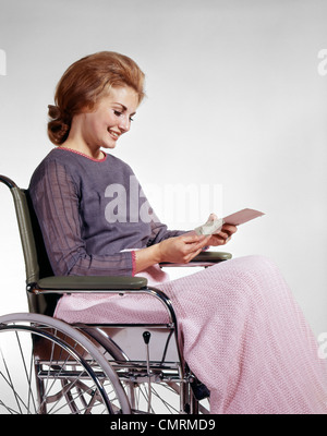 Anni sessanta donna sorridente HEALTHCARE paziente seduto nella sedia a rotelle ASSICURAZIONE DI LETTURA LA LETTERA DI CONTROLLO Foto Stock