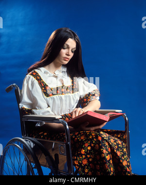 1970 anni settanta retrò donna capelli lunghi marrone seduto nella sedia a rotelle libro lettura Patient Healthcare Foto Stock