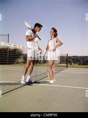 1960 anni sessanta retrò sorridente giovane uomo donna in piedi su lati opposti del NET CAMPO DA TENNIS HOLDING RACCHETTE RICREAZIONE Foto Stock