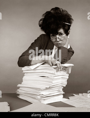 Anni sessanta anni settanta donna Segretario matita BLOCCATO IN TAQUINÉ capelli espressione oberato di lavoro inclinazione pila di documenti cartacei Foto Stock