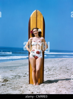 Negli anni quaranta anni cinquanta donna sorridente indossando Floral stampare BIKINI COSTUME DA BAGNO & fiori nei capelli appoggiata contro la tavola da surf guardando la fotocamera Foto Stock