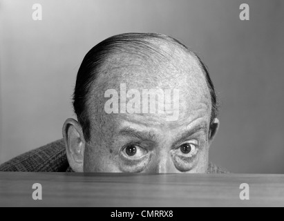 Anni sessanta BALDING MAN PEEKING SU DESKTOP CON solo la metà superiore della testa mostra guardando la fotocamera Foto Stock