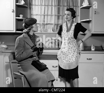 Trenta anni quaranta due donne SEDUTA IN CUCINA a parlare una casalinga casalinga in una catenaria in cappotto e HAT visitando insieme Gossip Foto Stock