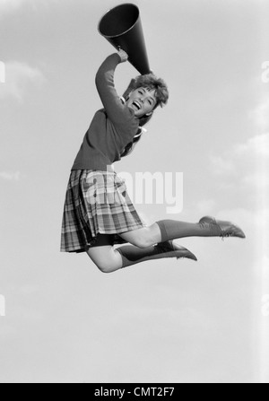 Anni sessanta ragazza adolescente cheerleader tifo saltare in aria tenendo megafono guardando la fotocamera Foto Stock