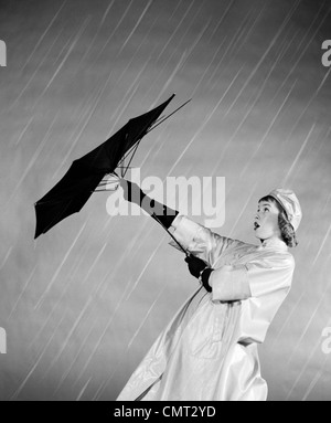 Anni Cinquanta la donna in cappotto di pioggia cercando di utilizzare ombrello rotto nel vento Foto Stock