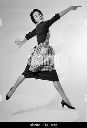 Anni Cinquanta donna casalinga nel grembiule GINGHAM correre e saltare in aria sorridente e puntare il dito indice STUDIO tacco alto scarpe Foto Stock