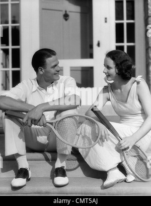 1930s GIOVANE UOMO DONNA NEL TENNIS WHITES HOLDING racchette seduta parlando sui gradini davanti la casa all'aperto Foto Stock