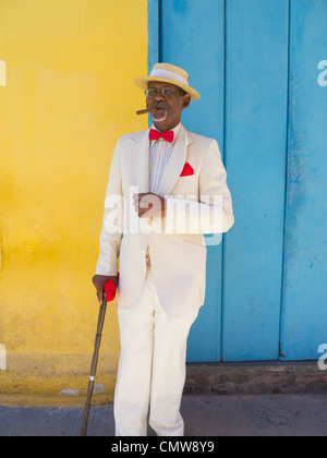 Un buio anziano gentiluomo cubano in un diportista più paglia hat, abito bianco, rosso il filtro bow tie, e un sigaro si erge da una parete colorata a l'Avana. Foto Stock