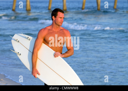 Atletica Giovane maschio adulto surfer porta la sua tavola da surf lungo la spiaggia con palificazioni di un molo in background. Foto Stock