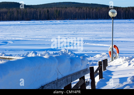 Coperta di neve staccionata in legno e un lampione con salvagente a coperta di neve lago ghiacciato in inverno Foto Stock