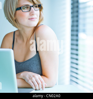 Ritratto di una giovane donna pensively guardando fuori della finestra mentre è seduto davanti al suo computer portatile Foto Stock