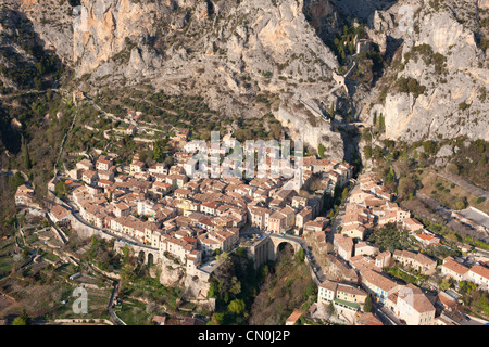 VISTA AEREA. Borgo medievale ai piedi di una massiccia scogliera. Moustiers-Sainte-Marie, Alpes-de-Haute-Provence, Francia. Foto Stock