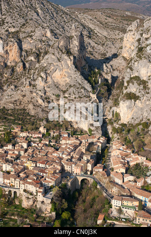 VISTA AEREA. Borgo medievale ai piedi di una massiccia scogliera. Moustiers-Sainte-Marie, Alpes-de-Haute-Provence, Francia. Foto Stock