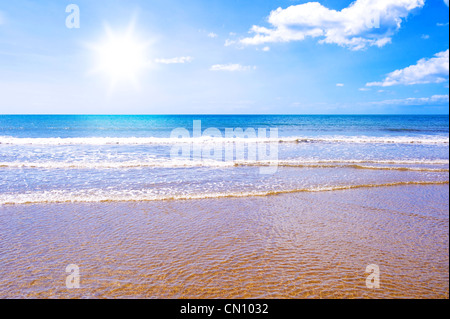 Foto di onde che si infrangono sulla spiaggia dorata con il sole che splende in un cielo blu. Foto Stock