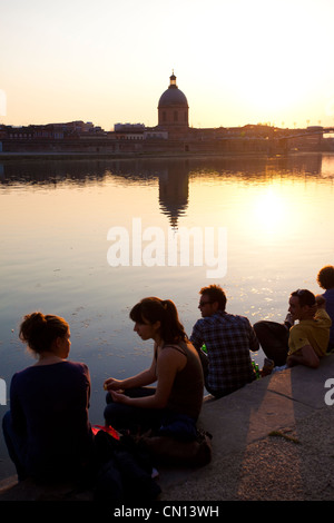 Persone sedersi sulle rive di La Garonne come il sole tramonta chiudere dietro la cupola de la tomba, Toulouse, Francia Foto Stock