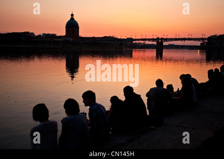 Persone sedersi sulle rive di La Garonna al tramonto con la vista della Cupola de la tomba e St Pierre bridge, Toulouse, Francia Foto Stock