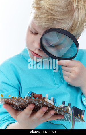 Piccolo ragazzo biondo guardando una scheda madre attraverso la lente di ingrandimento Foto Stock