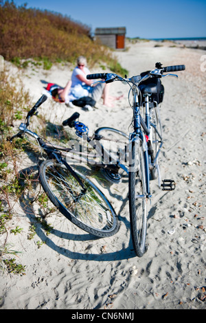 Due signore biciclette sul West Wittering beach, West Sussex, con la signora ciclisti fuori fuoco in appoggio in background Foto Stock