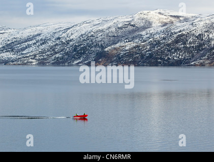 Un rosso barca con 2 persone che si spostano da sinistra a destra su un fiordo con montagne innevate dietro Foto Stock