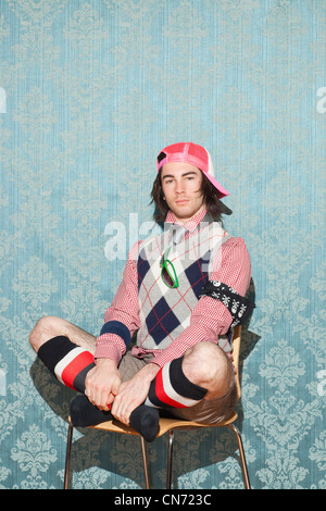 Ritratto di giovane caucasico uomo che indossa cappello rosa seduti su una sedia, gambe piegate con sfondo. Foto Stock